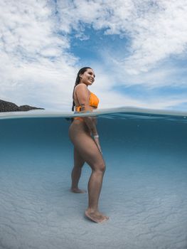 A woman in an orange bikini smiles while standing half submerged in clear ocean water.