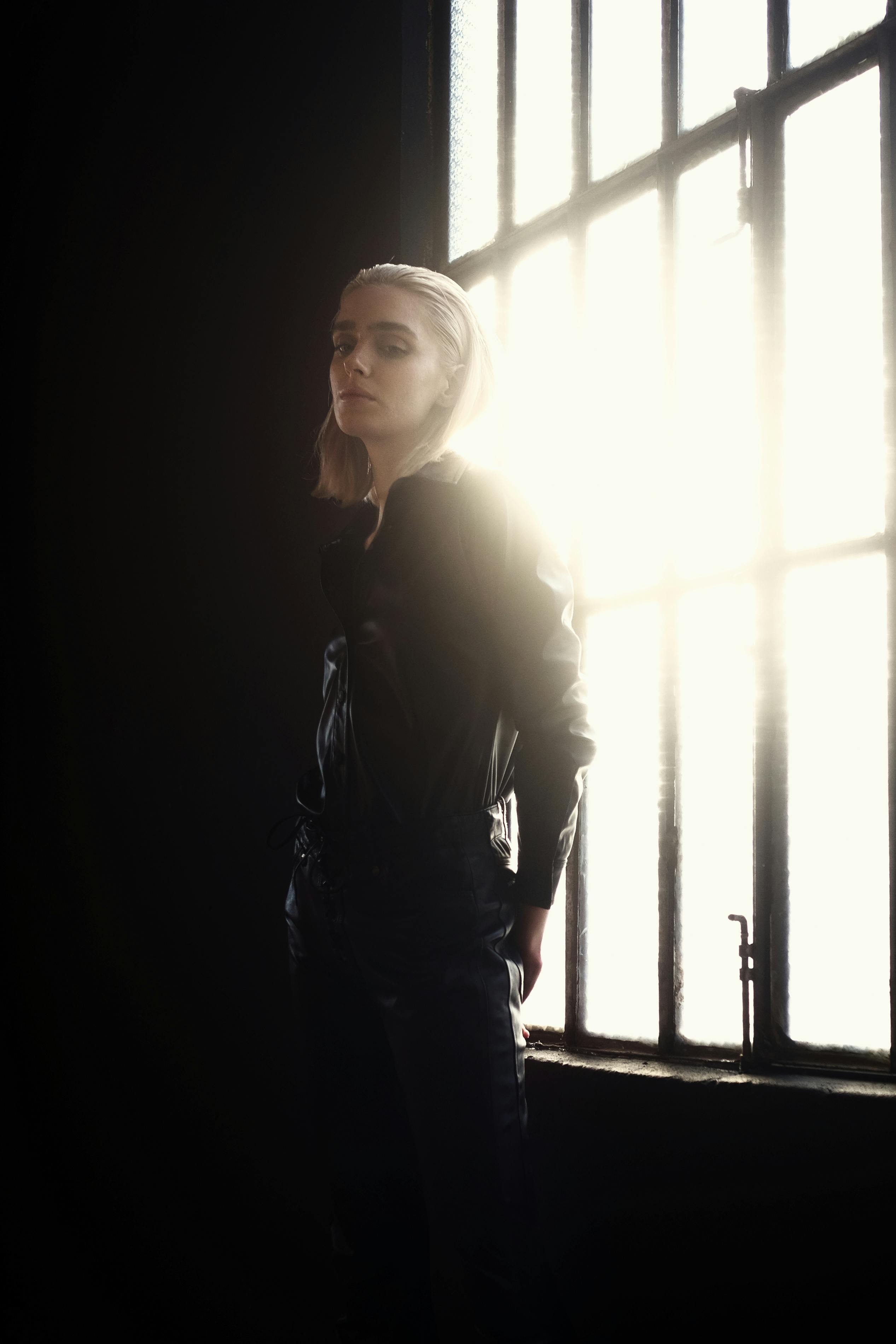 A dramatic portrait of a woman in leather, standing by a window with backlighting.