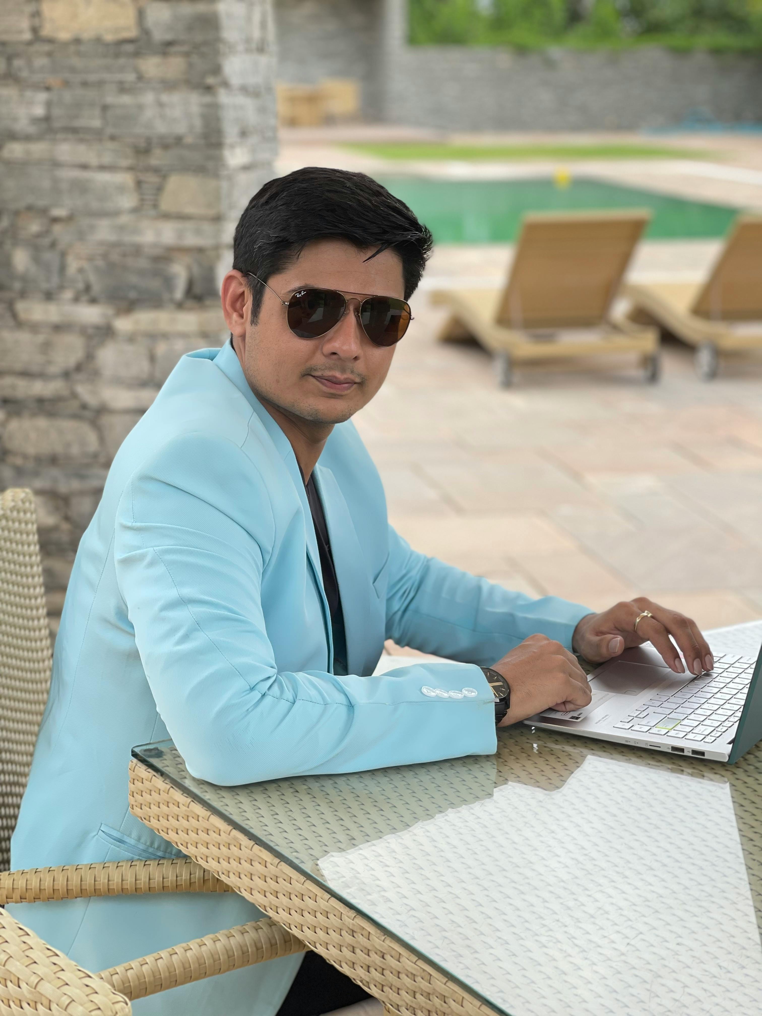 Man in sunglasses working on a laptop by the pool, dressed in a stylish blue blazer.