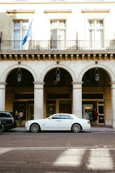 White luxury car parked beside elegant building facade with arched design, capturing a sophisticated urban scene.