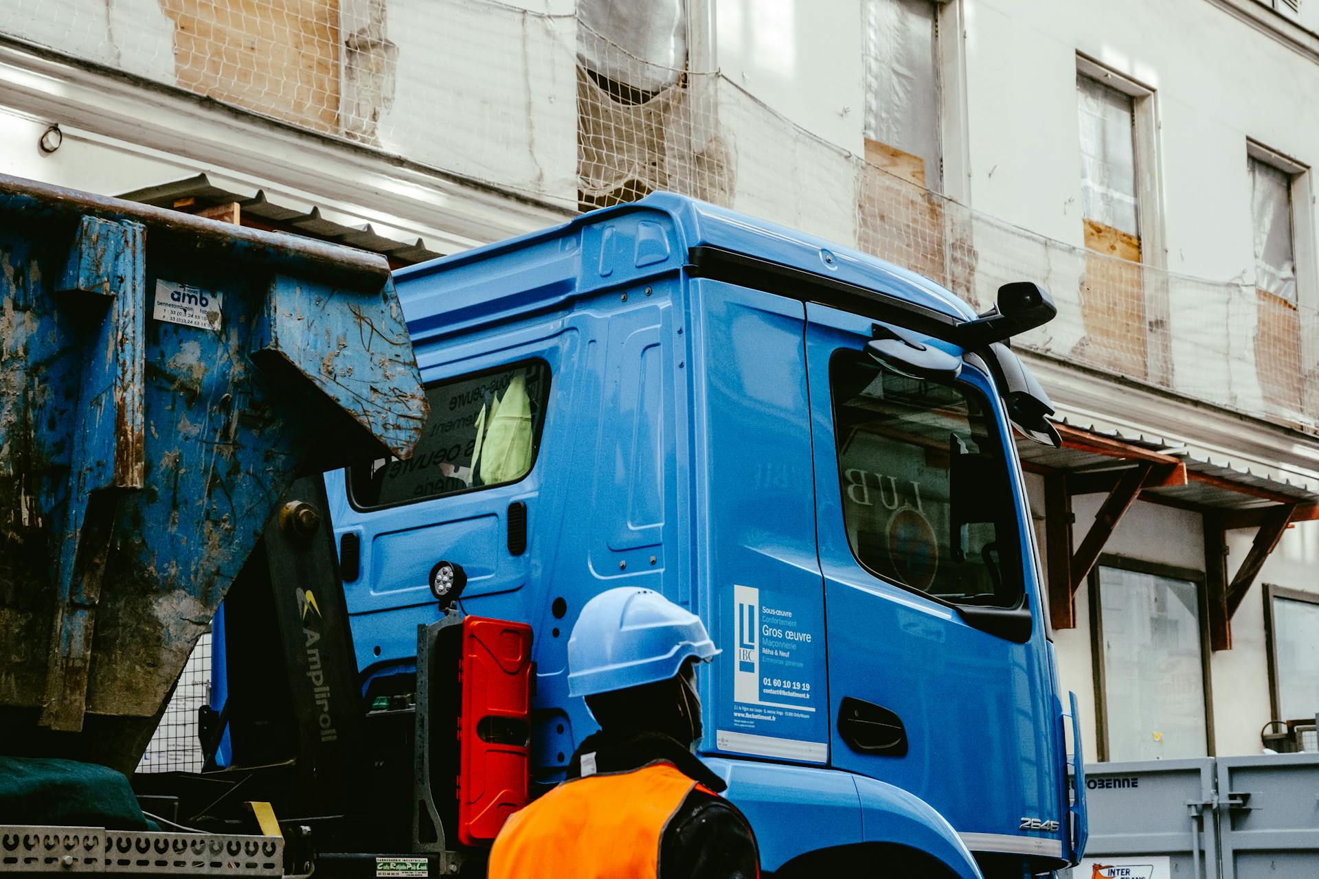 Construction worker standing by a blue truck at an urban building site, wearing a safety vest and hard hat.
