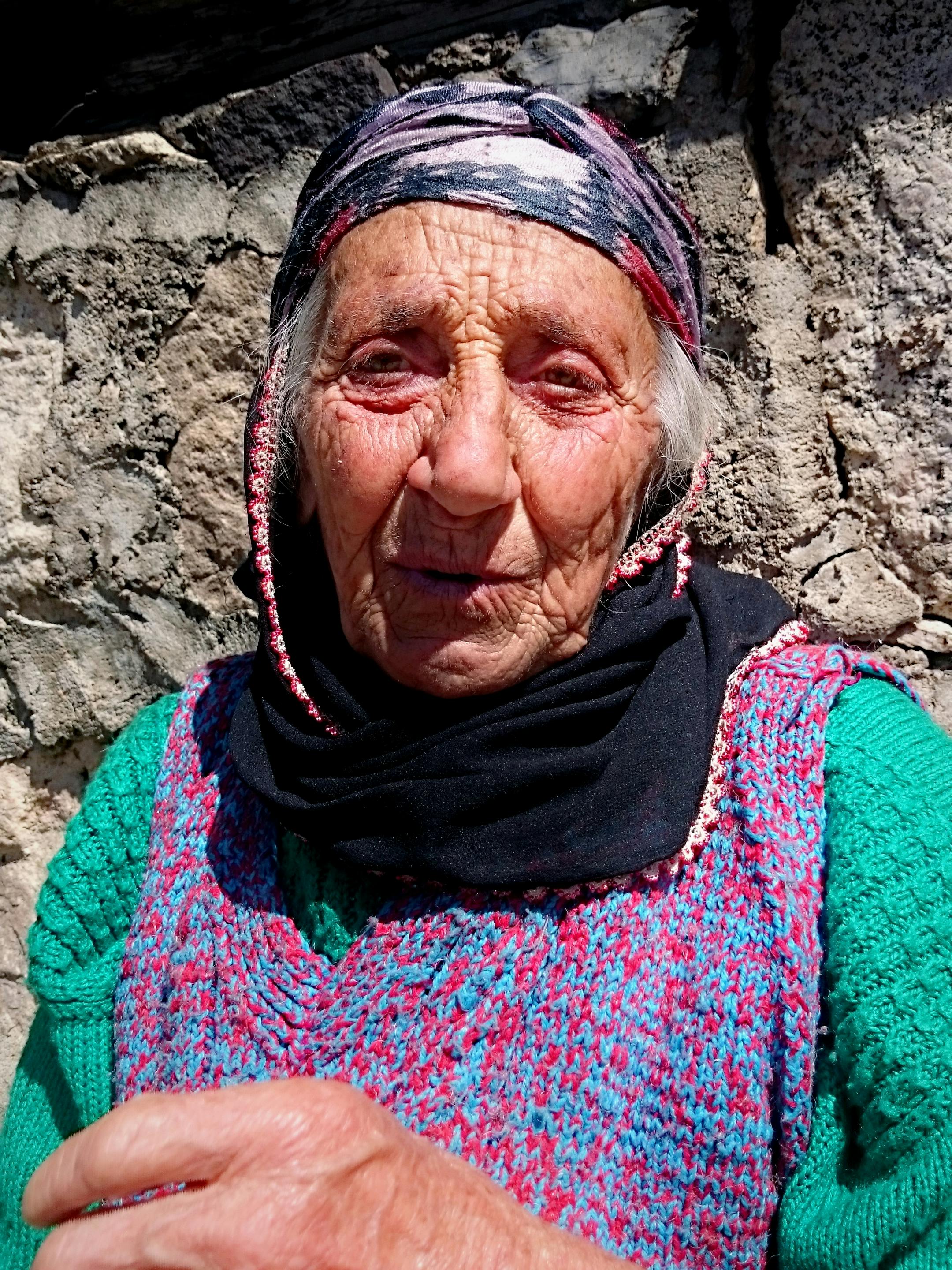 Close-up shot of a senior woman with a colorful headscarf and sweater against a stone wall on a sunny day.