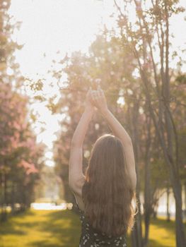 Back view of a woman raising arms in a sunlit park surrounded by trees.