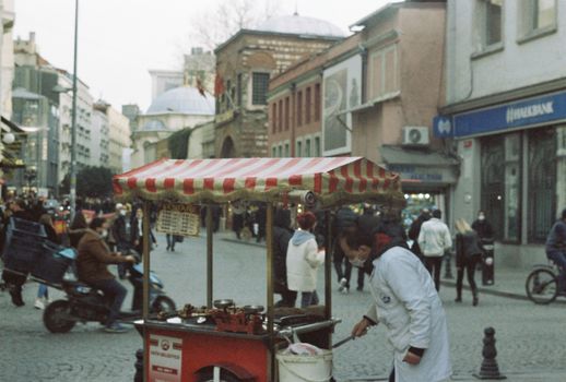 Busy street scene with a vendor at a food stall and people walking by. Urban atmosphere.
