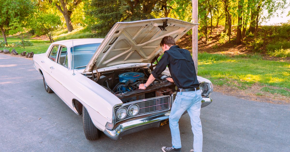 A man leaning under the open hood of a white classic car on a sunny tree-lined road