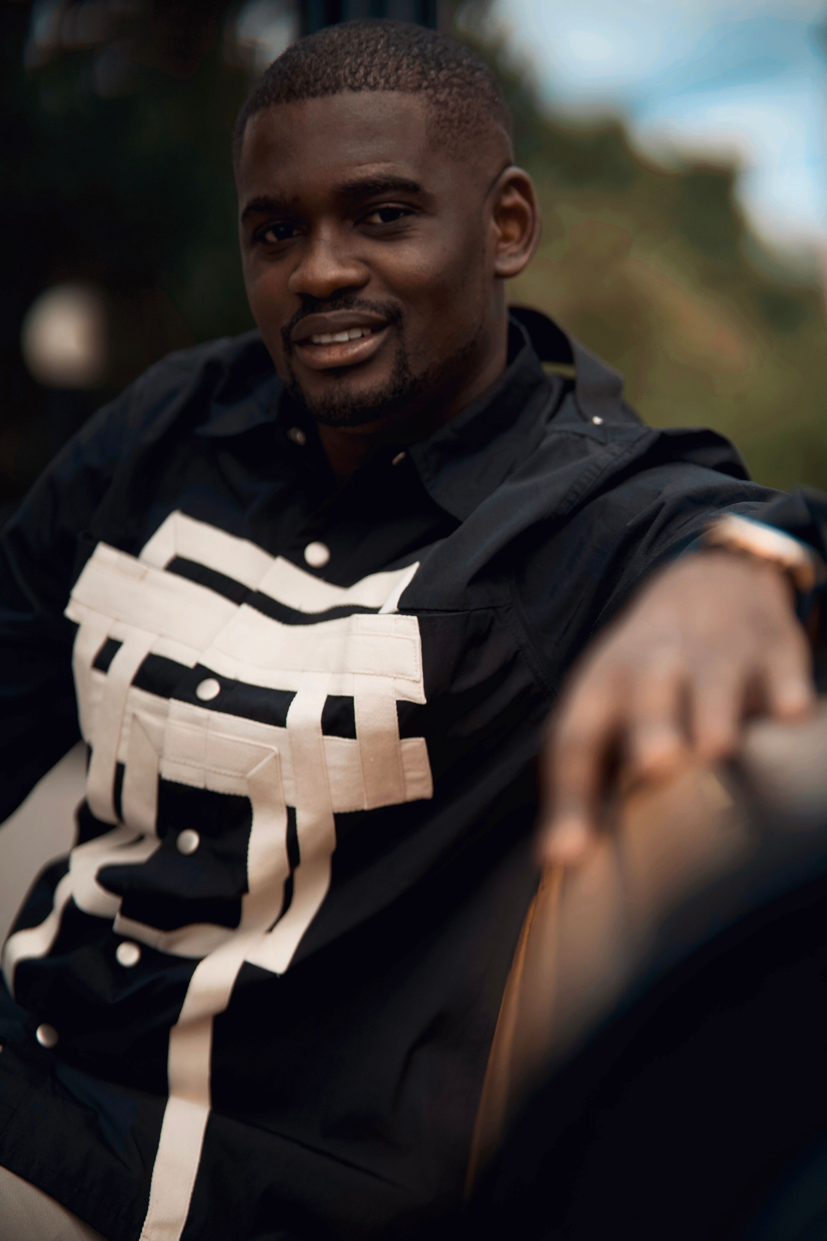 African American man smiling in a black printed shirt, sitting outdoors.