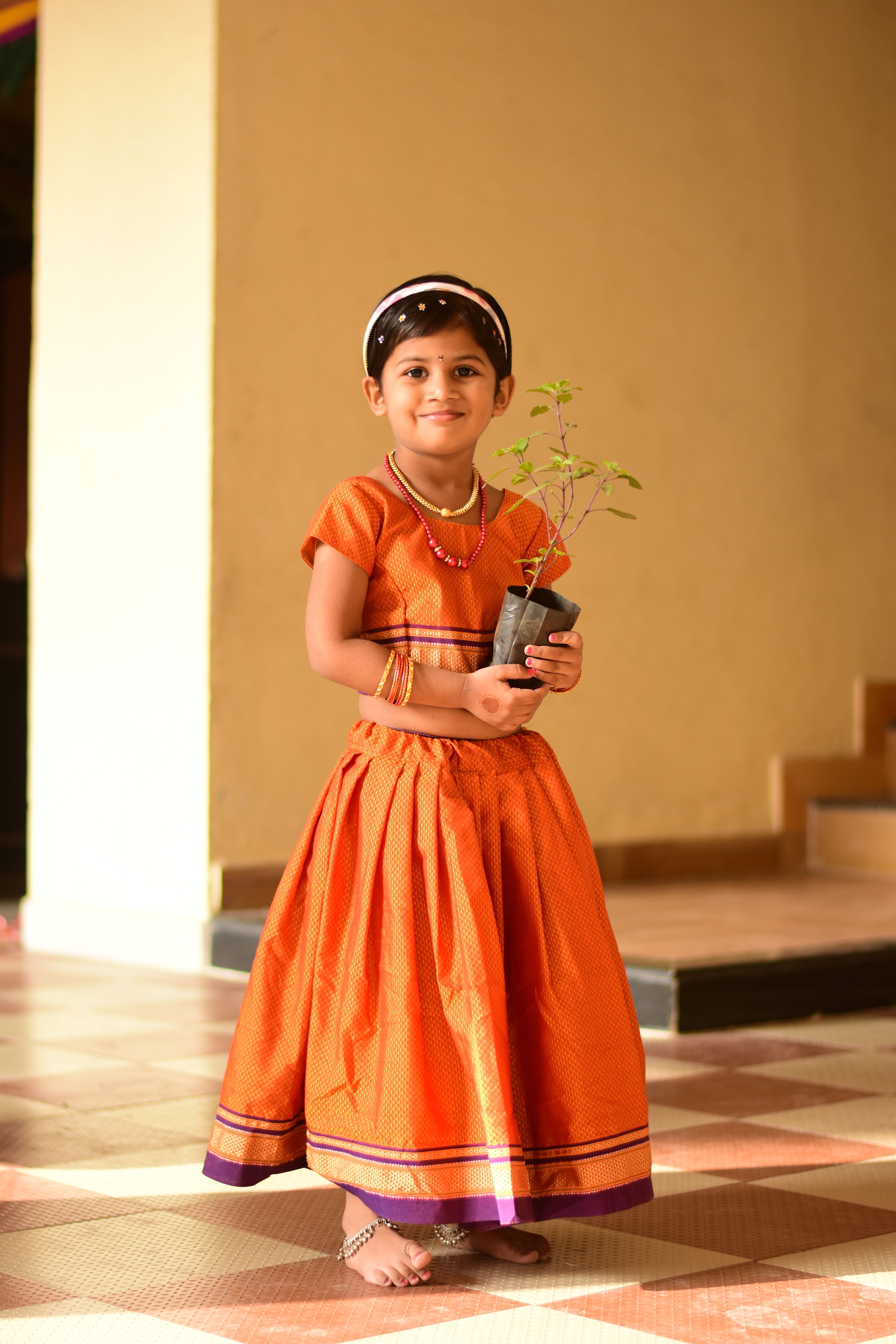A smiling South Asian girl in an orange dress holds a potted plant indoors.