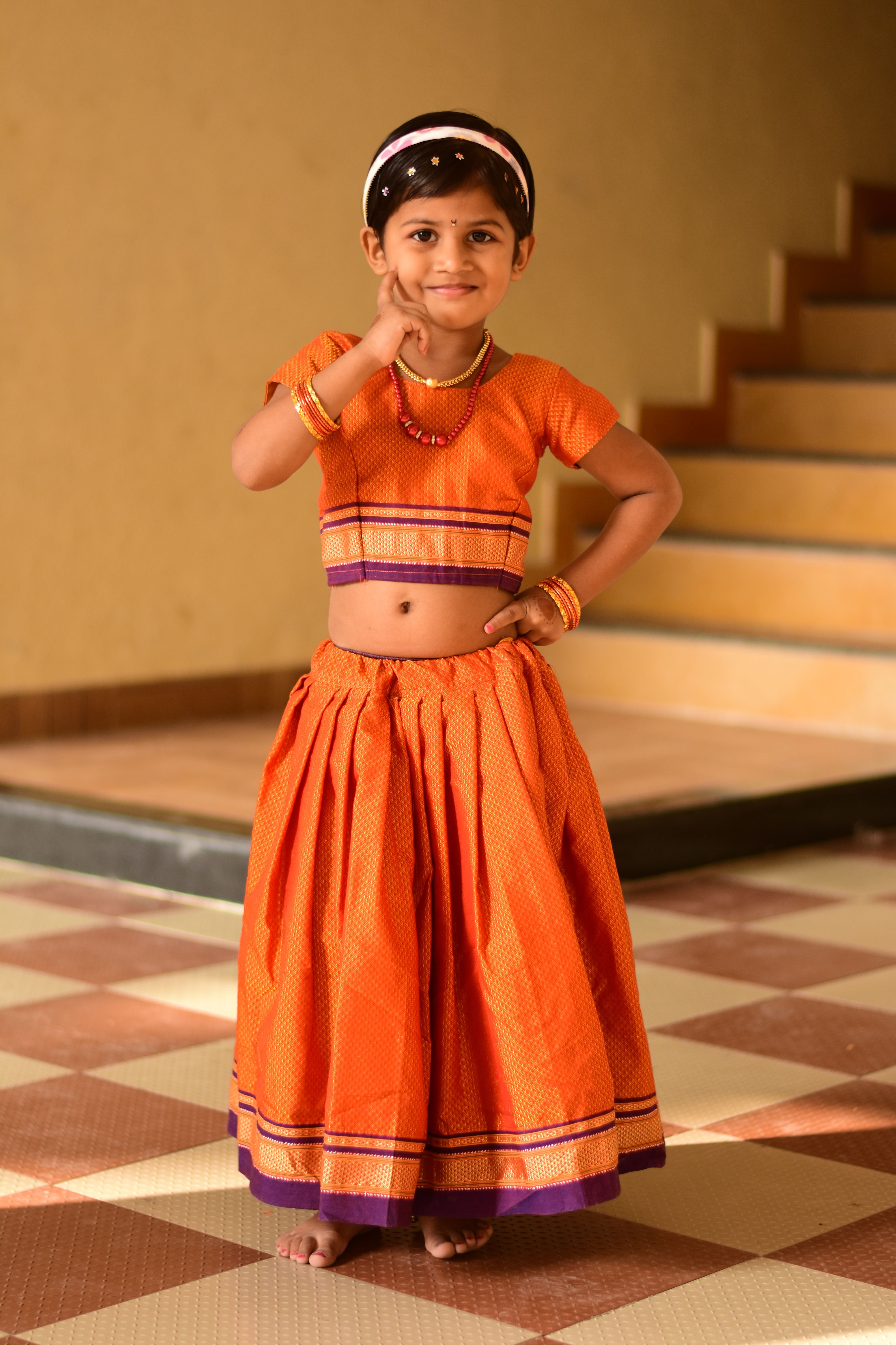 Smiling girl in an orange dress poses indoors, showing traditional Indian attire.