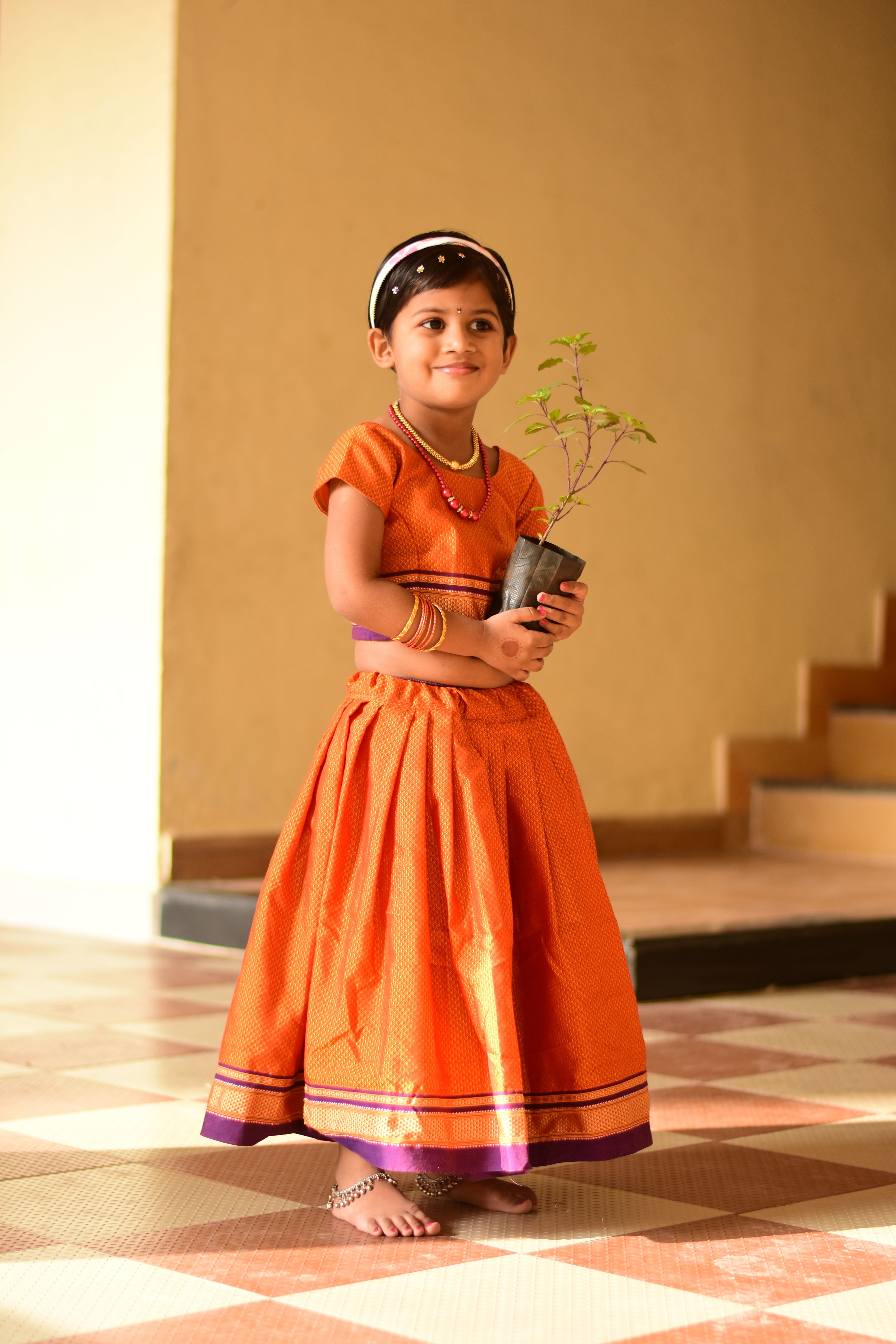 A smiling young girl in orange ethnic attire holding a potted plant indoors.