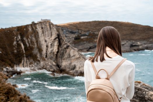 Woman in white cardigan with backpack observing rocky coastline and ocean.