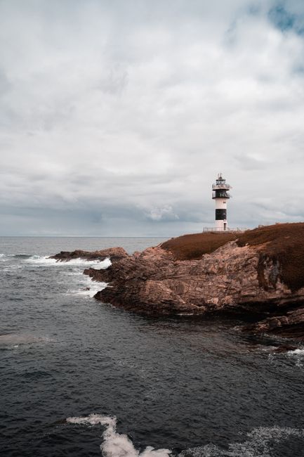 Lighthouse standing on a rocky coastal cliff with waves crashing below under an overcast sky.
