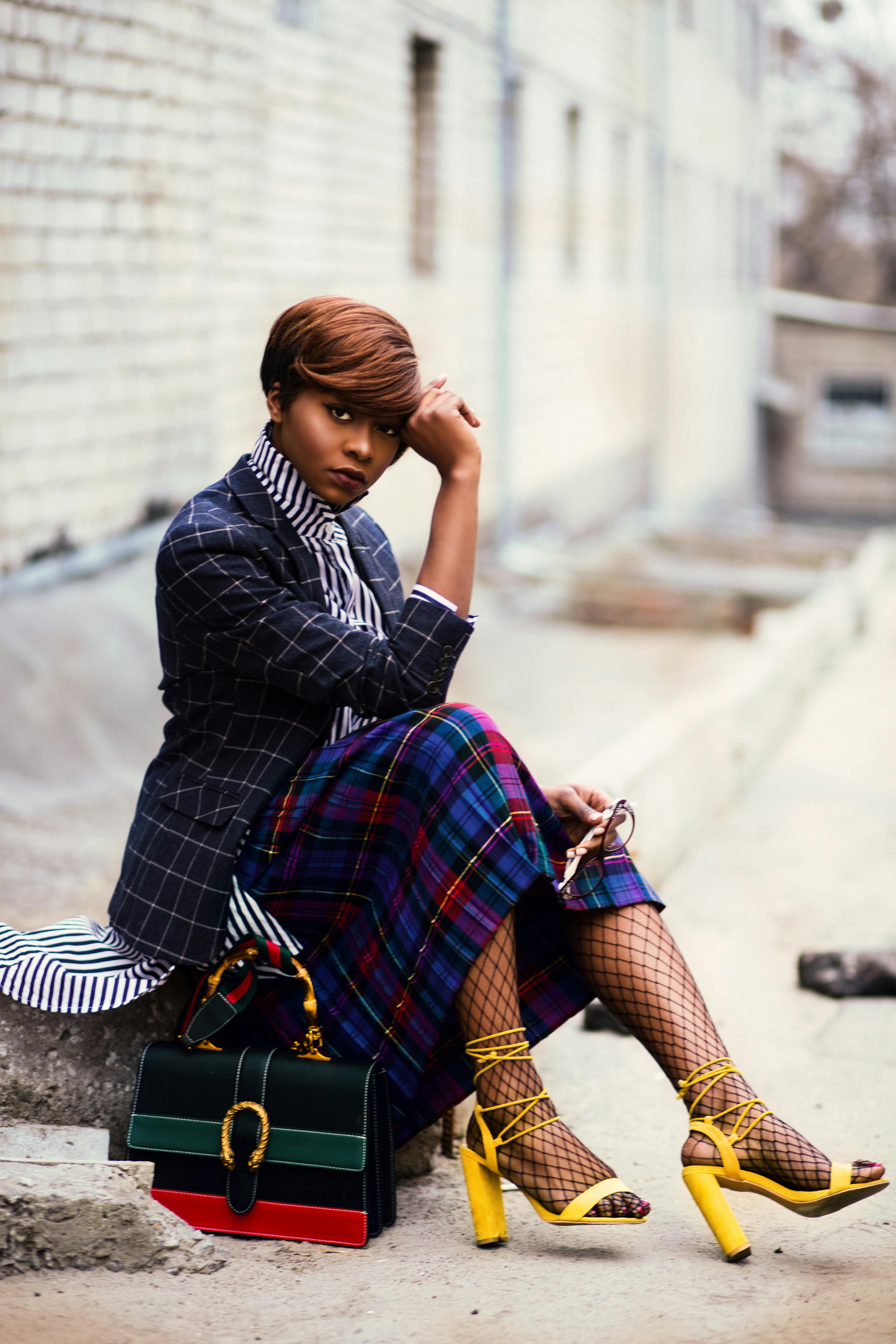 woman wearing black and grey tattersall blazer and multicolored plaid skirt with black mesh stocking and yellow chunky heeled sandals sitting on grey concrete pathway