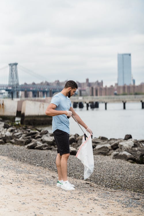 Man in White T-shirt and Black Shorts Holding White Plastic Bag Standing on Gray Rock