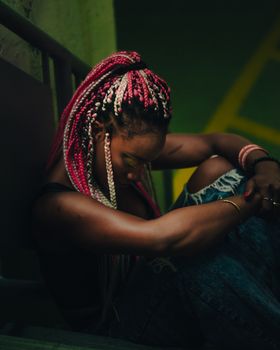A woman with vibrant pink dreadlocks sits thoughtfully indoors, reflecting a moody atmosphere.