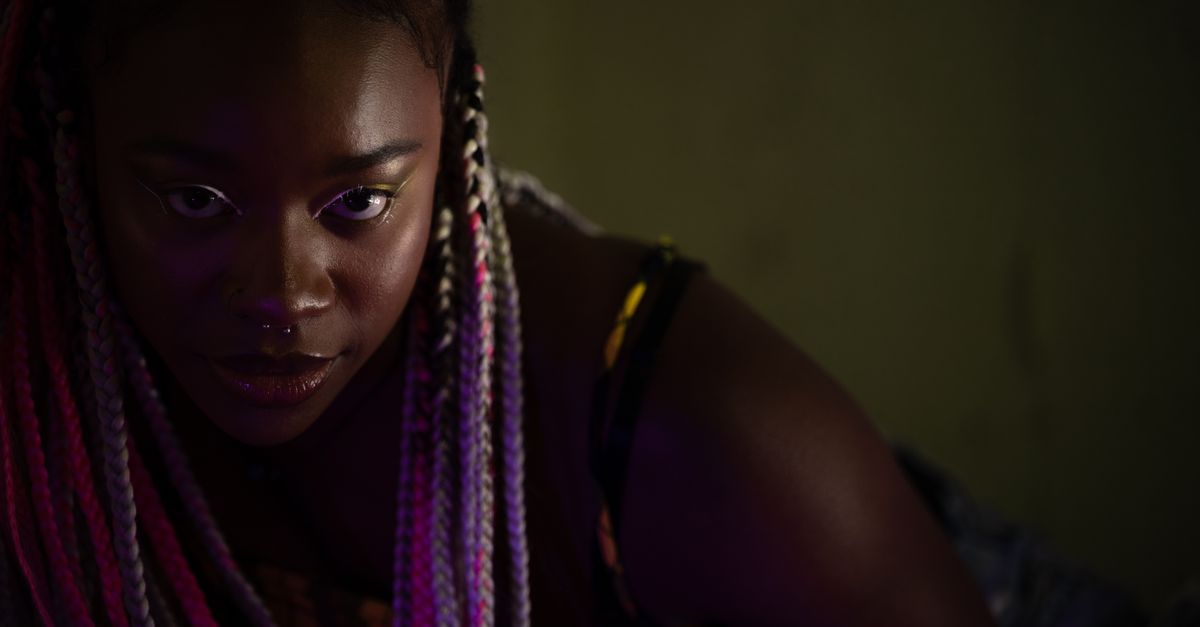 Retrato close-up dramático de uma mulher com dreads vibrantes e piercing no nariz.