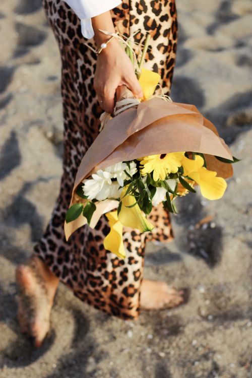 Free A woman walks barefoot on a sandy beach, holding a vibrant bouquet of flowers. Stock Photo