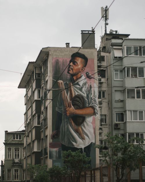 Free A large mural depicting a man with a bird on an urban building's exterior, surrounded by trees. Stock Photo