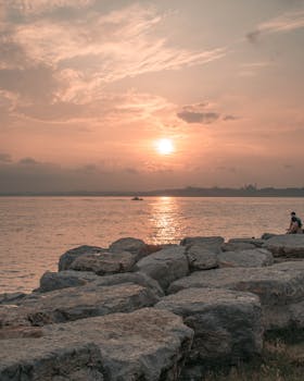 Tranquil sunrise over the sea with a rocky coastline, reflecting warm pink hues.