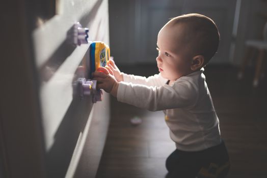 A cute baby exploring magnetic toys on a cupboard, illuminated by natural light indoors.