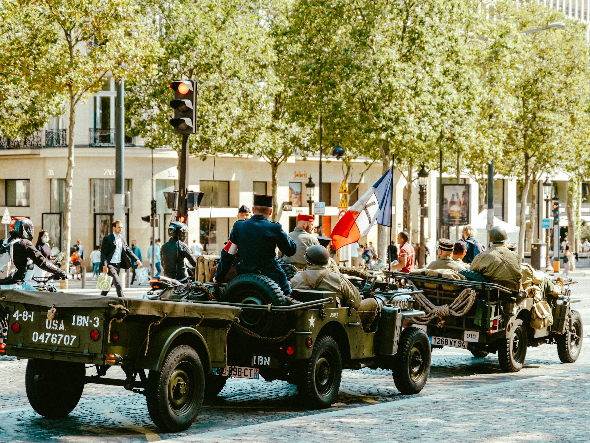 Vintage military jeeps in a parade, featuring soldiers and French flags in an urban setting.