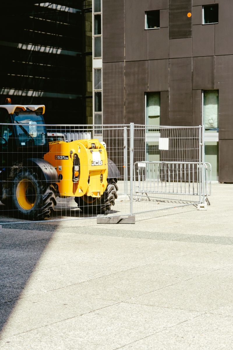 A yellow tractor parked behind a metal fence at an urban construction site.