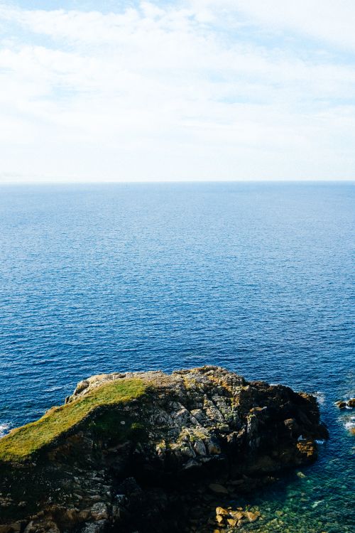 Free A scenic view of a rocky coastline with a vibrant blue sea under a cloudy sky. Stock Photo