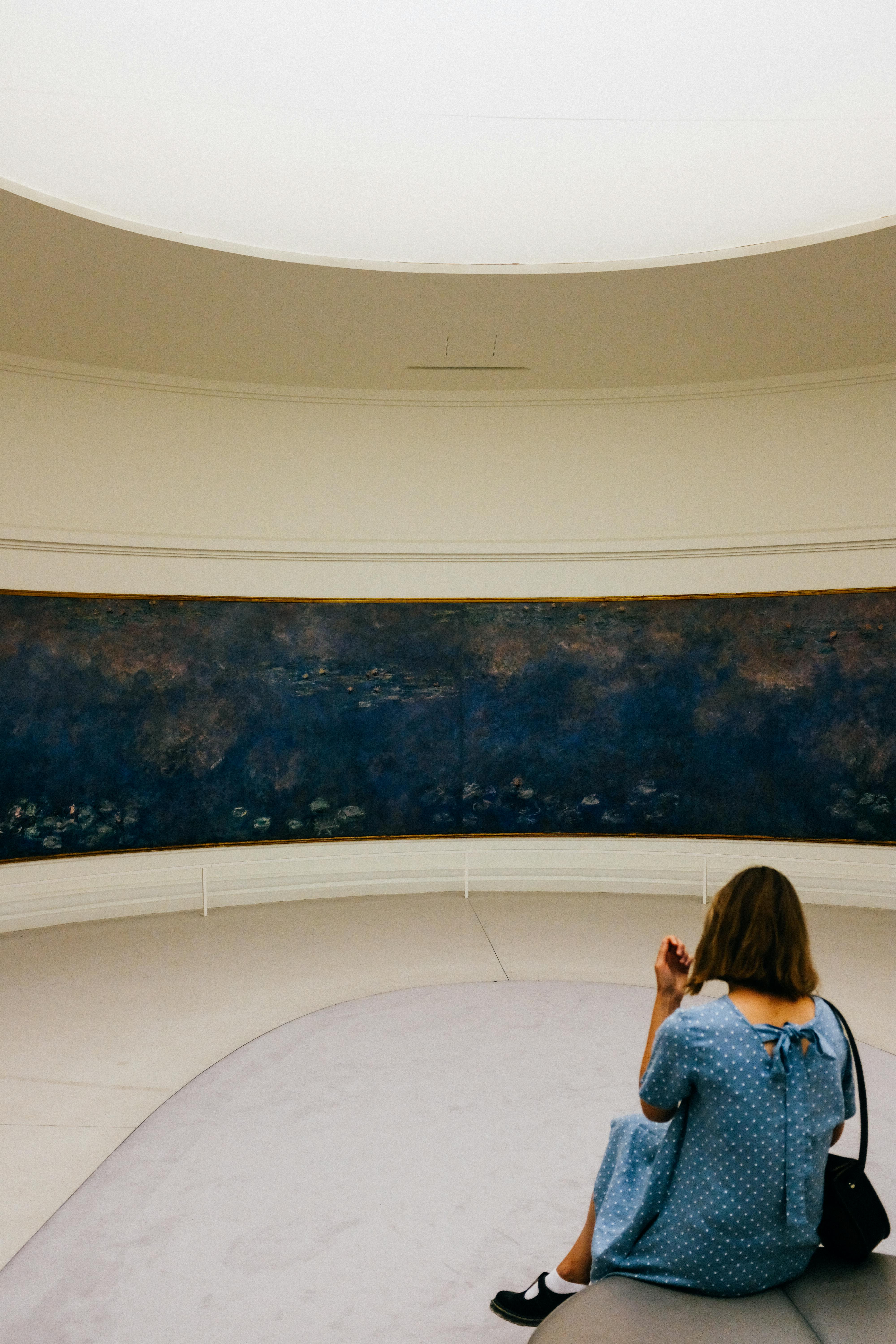 A woman in a blue dress sits in a museum, admiring a large painting in a serene atmosphere.