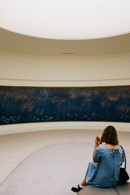 A woman in a blue dress sits in a museum, admiring a large painting in a serene atmosphere.
