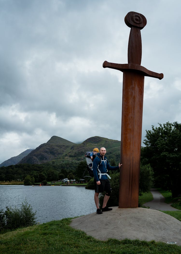 Man Standing Beside A Giant Sword Near A Body Of Water