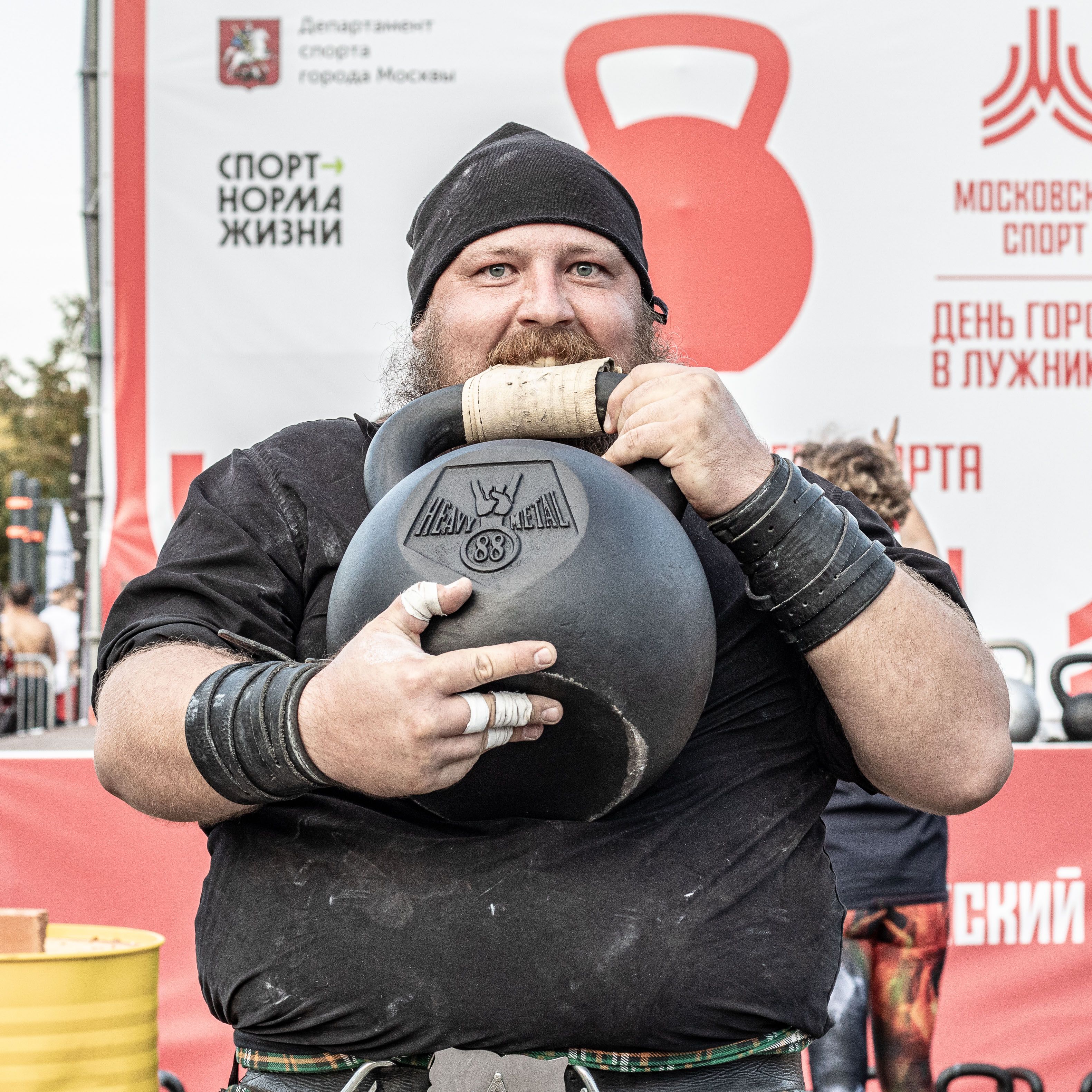 Close-up of a strongman enthusiast lifting a heavy kettlebell during an outdoor sports event.