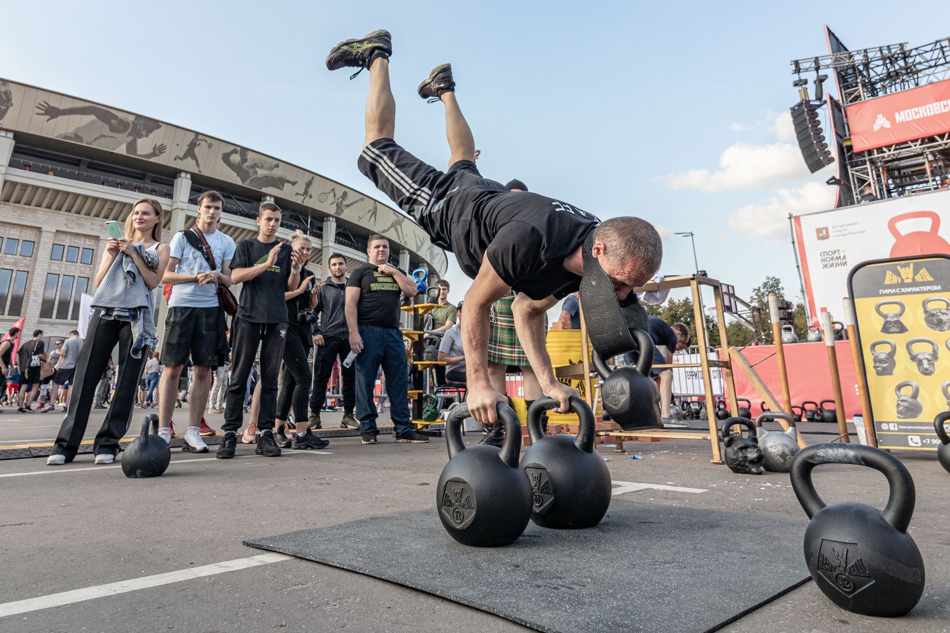 Man performing handstand on kettlebells during outdoor fitness event.