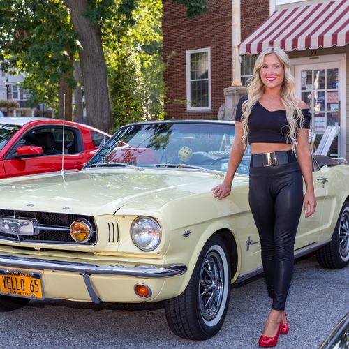 Free Blonde woman standing by a vintage yellow Mustang convertible at an outdoor car show. Stock Photo