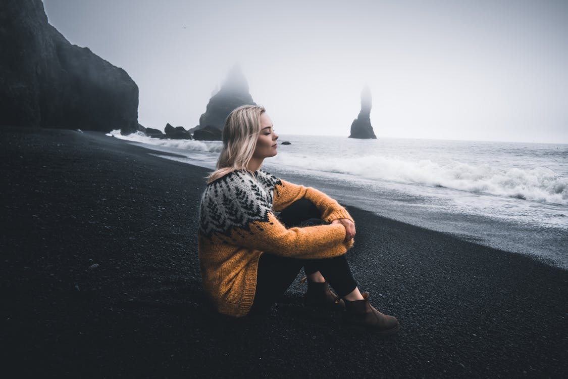 Woman in Black and White Long Sleeve Shirt and Brown Pants Sitting on Black Sand during