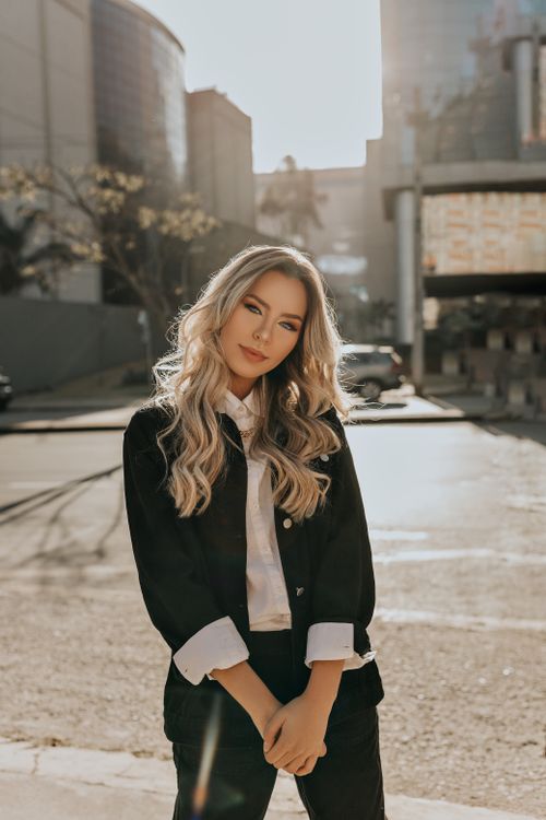 Free Fashionable woman with long wavy hair stands confidently in a sunlit cityscape. Stock Photo