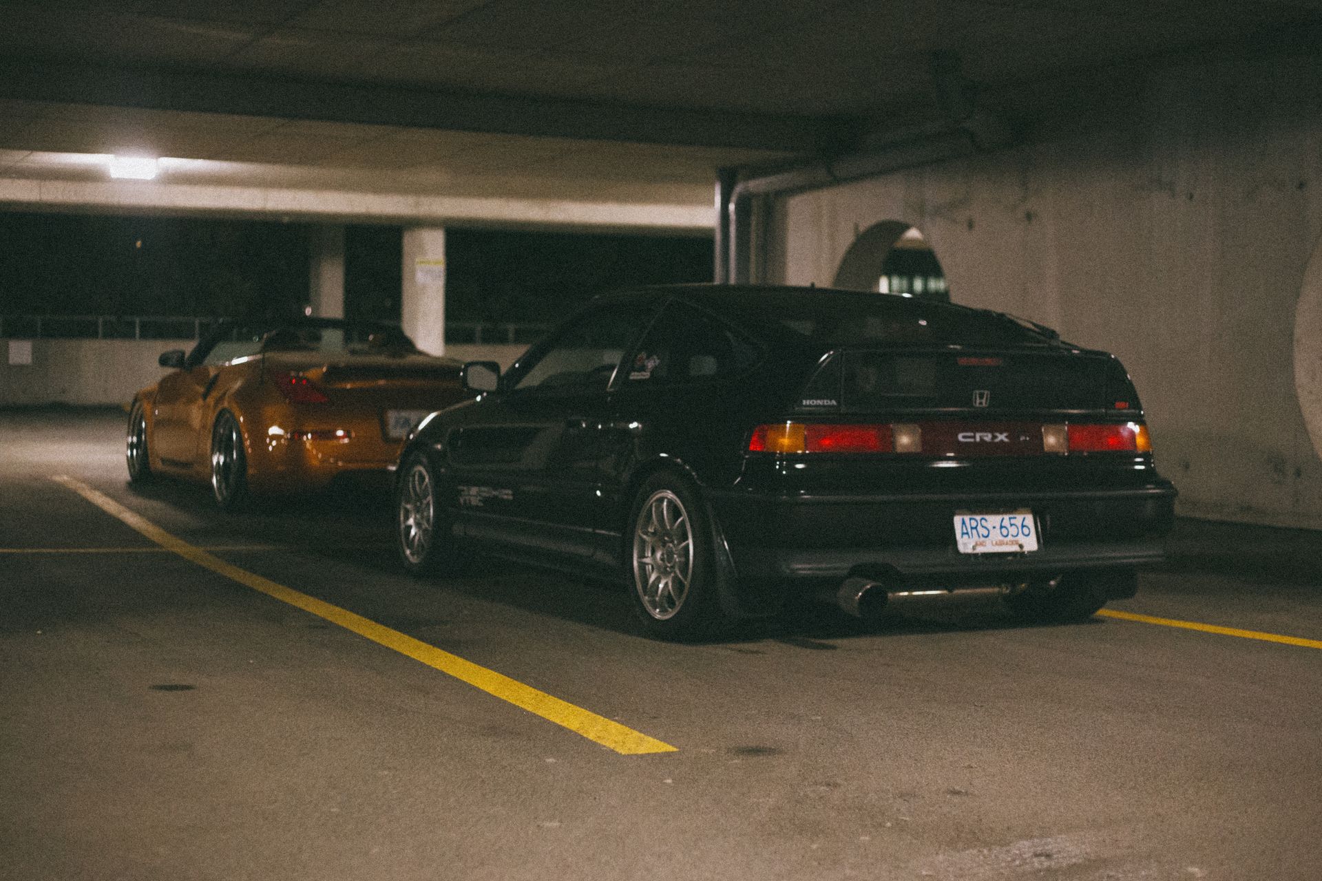 Black Honda CR-X and gold sports car parked in dimly lit garage.