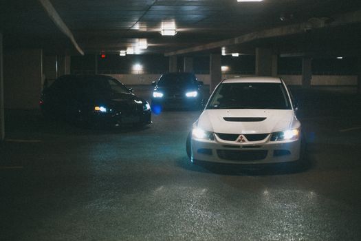 Automobiles with headlights on in a dimly lit underground parking lot.