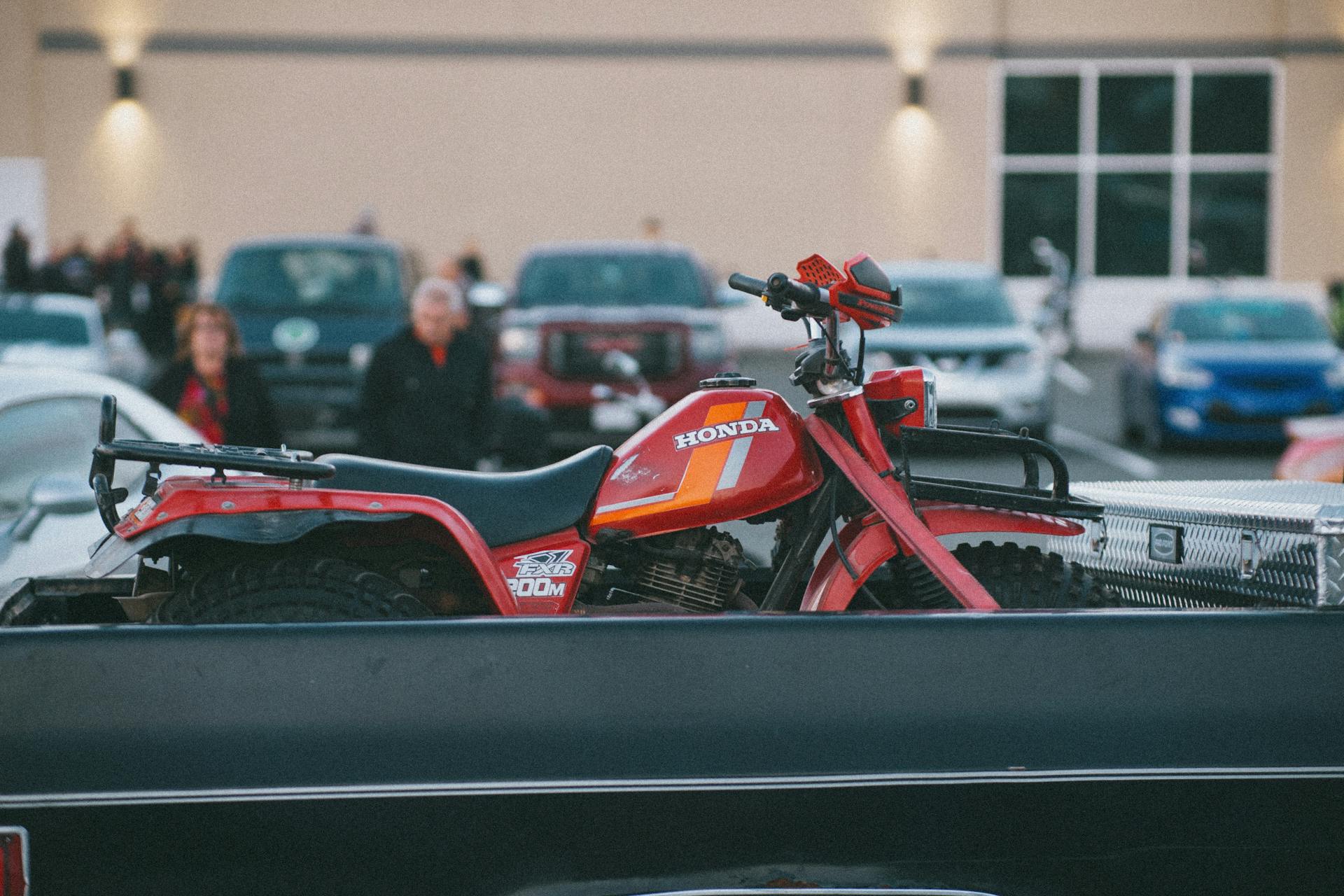 A red quadbike secured on a truck in a crowded parking lot.