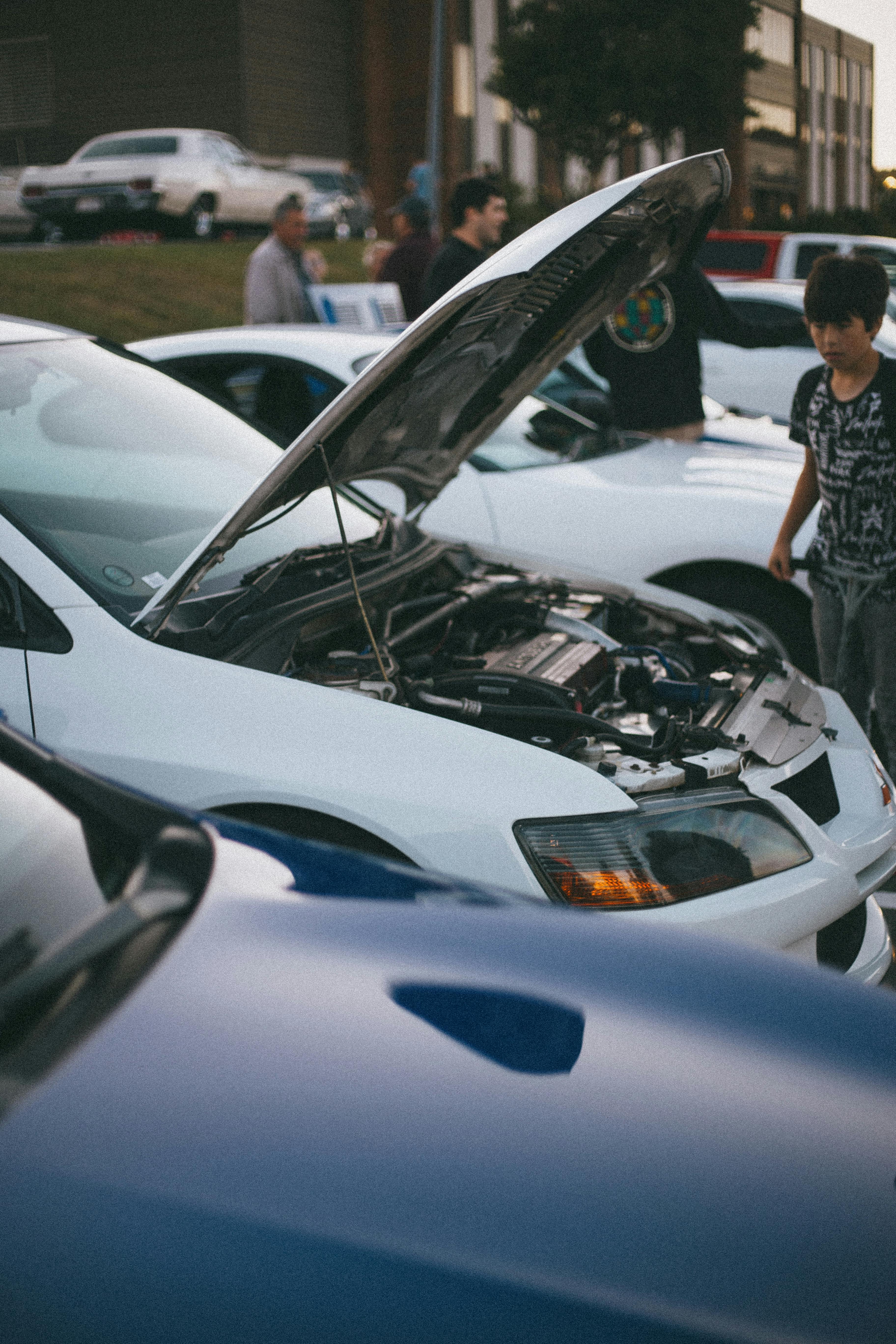 A white Mitsubishi Lancer Evolution with open hood at a car show, surrounded by people.