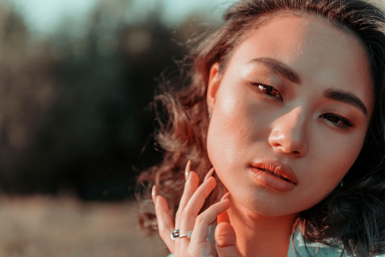 Woman with glowing skin lightly touches her cheek in warm outdoor golden light.
