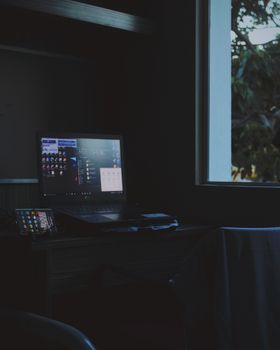 A dimly lit workspace featuring a laptop and smartphone on a wooden desk by a glass window.