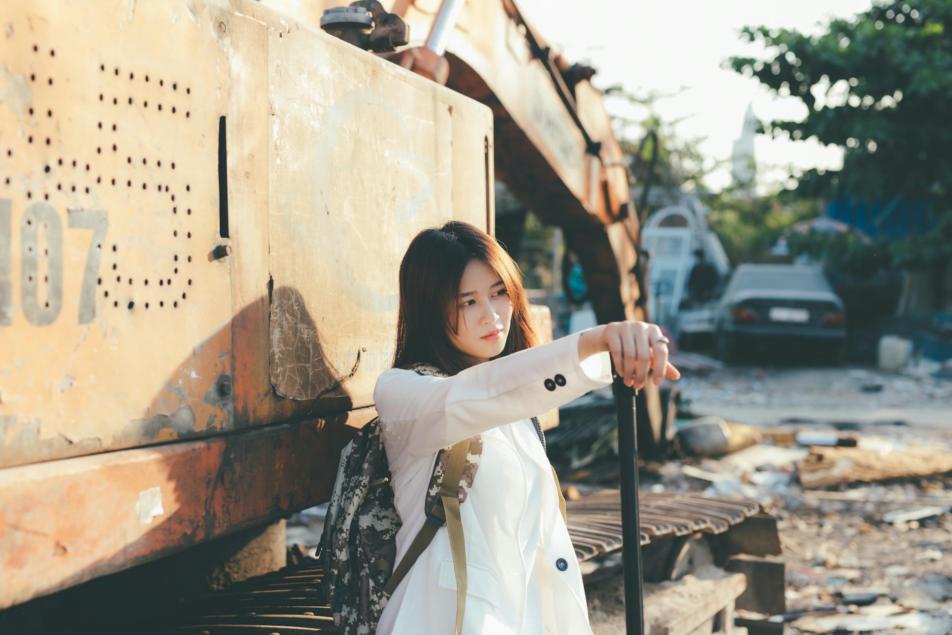 Young Asian woman in a white coat leaning on an excavator in an industrial outdoor setting.