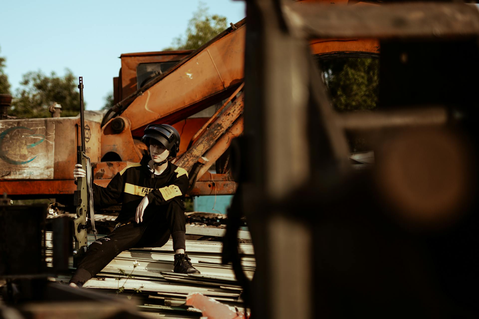 A person in tactical gear sitting beside an excavator in an outdoor setting.