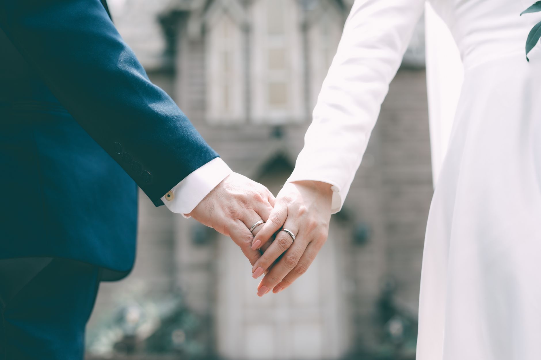 Close-up of a bride and groom holding hands, showcasing wedding rings and elegant attire.