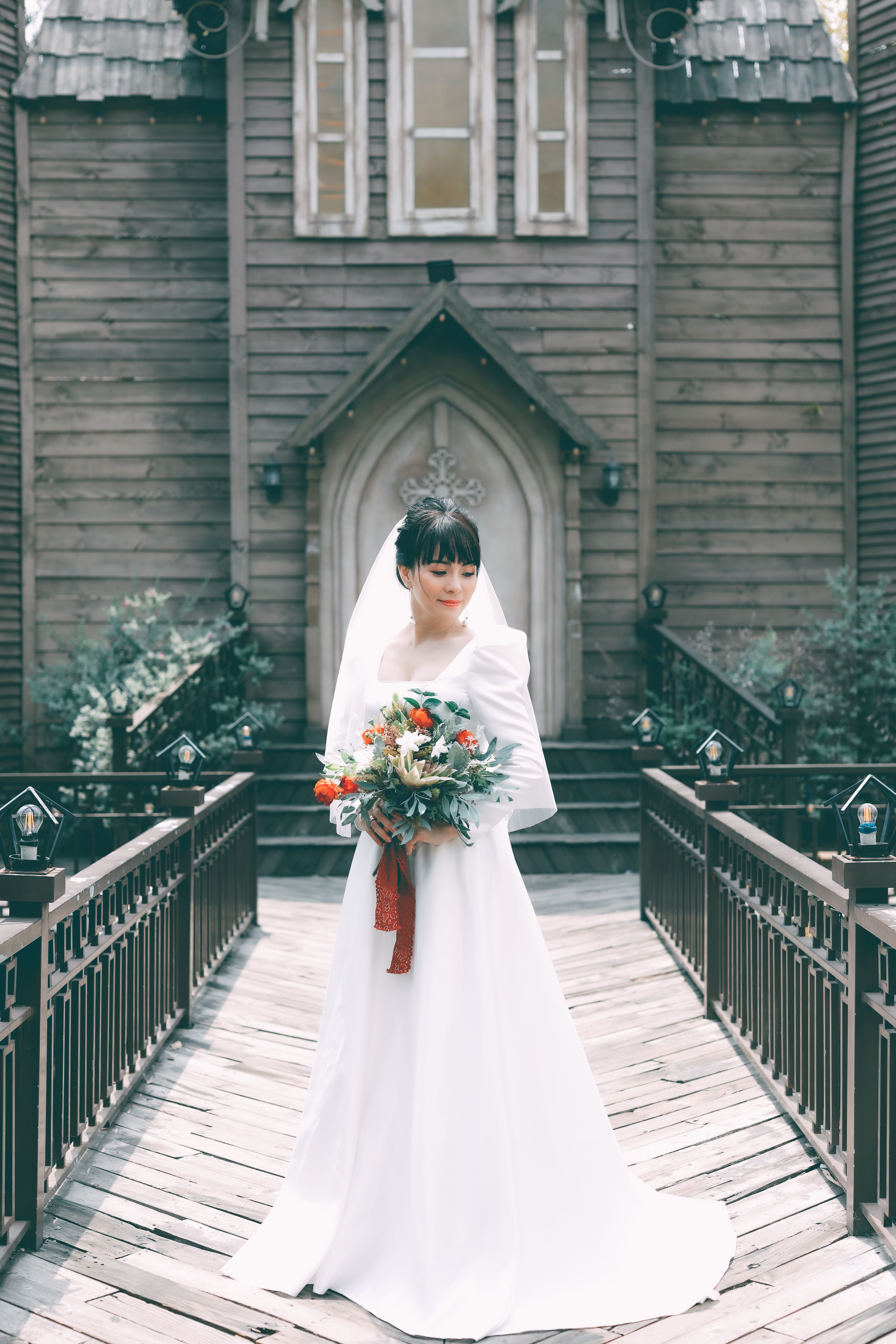 Beautiful bride in a white wedding dress standing outside a wooden chapel holding a colorful bouquet.