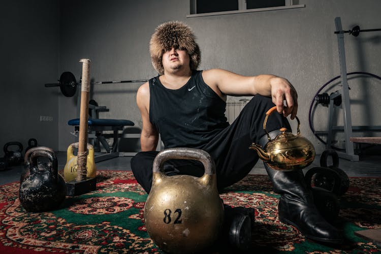 A Man In Black Clothes Sitting On The Carpet Beside The Kettle Bells