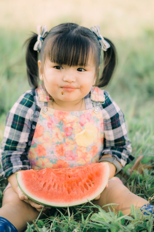 Free Cute child in floral outfit eating watermelon while sitting on grass, enjoying a summer day. Stock Photo