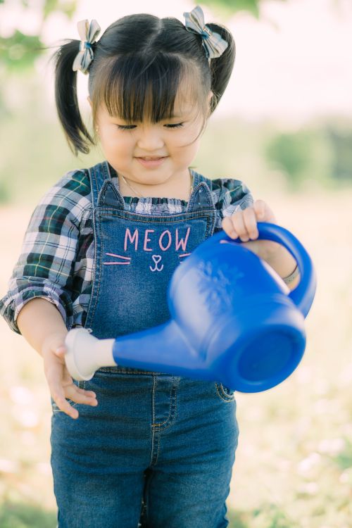 Free Cute child in overalls holding a blue watering can outdoors on a sunny day. Stock Photo