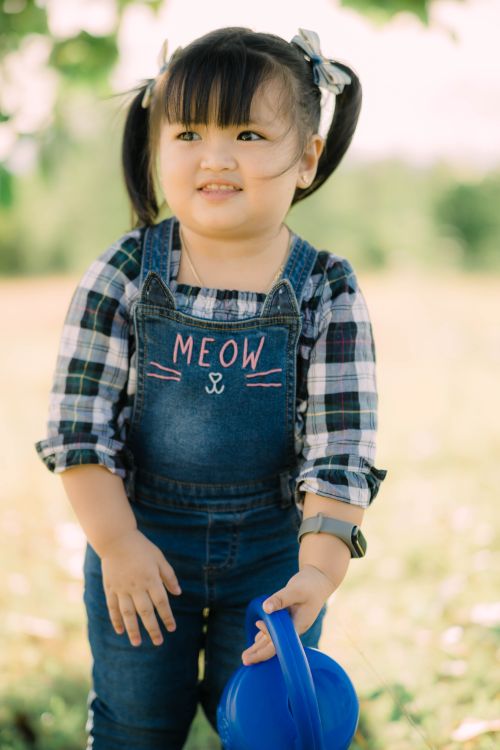 Free Adorable child in denim overalls holding a watering can in a sunny outdoor setting. Stock Photo