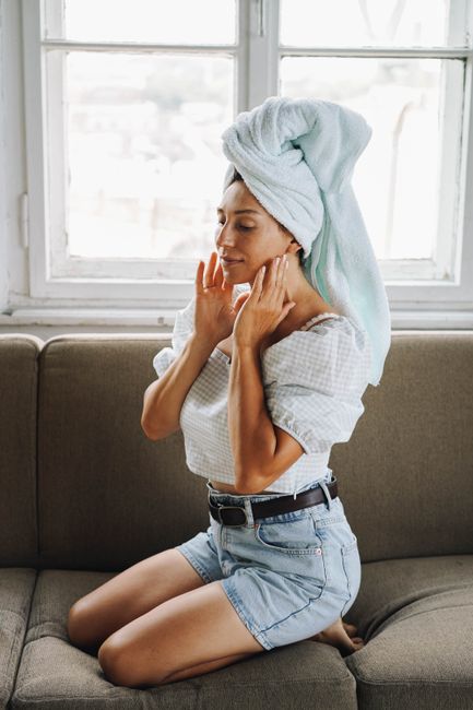 Young woman with a towel on her head kneeling and relaxing indoors by a window. Casual and serene setting.