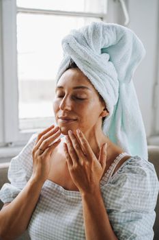 Woman with towel wrapped on hair enjoying a skincare routine by window.